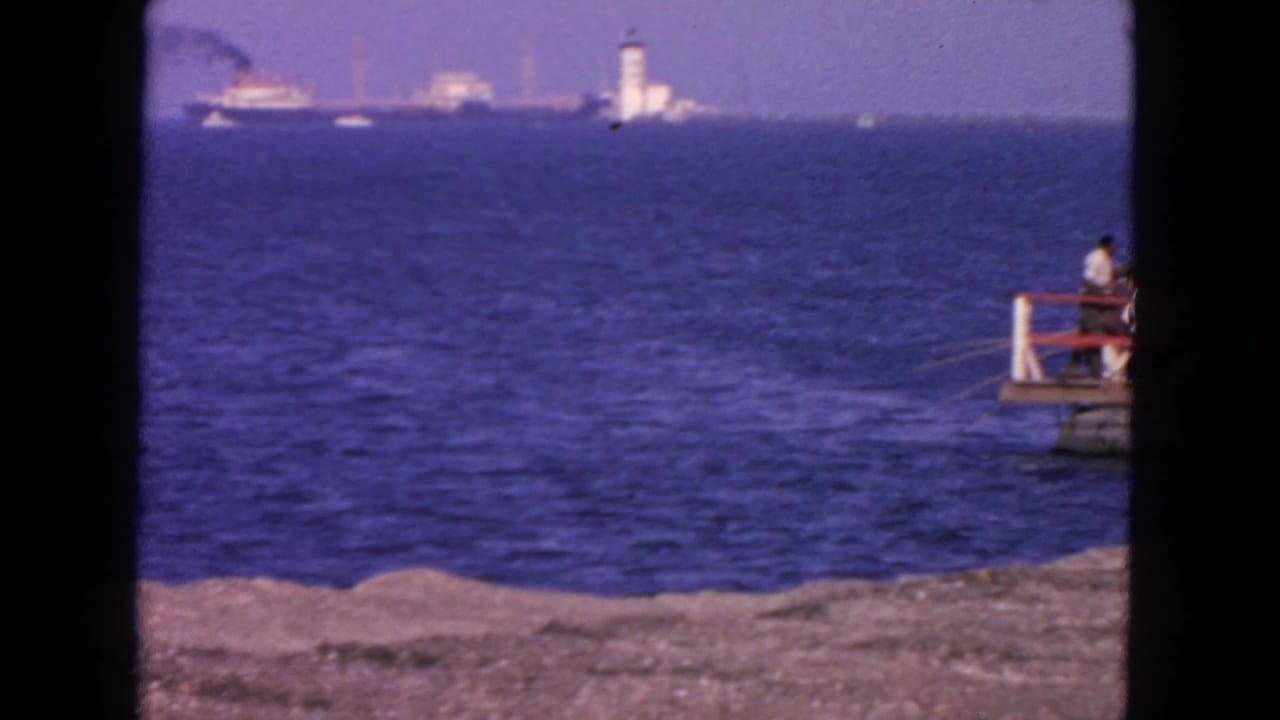 1963 San Francisco coastal view with blue water, distant ships, a white tower-like lighthouse, shoreline, and a person seated on a small pier.