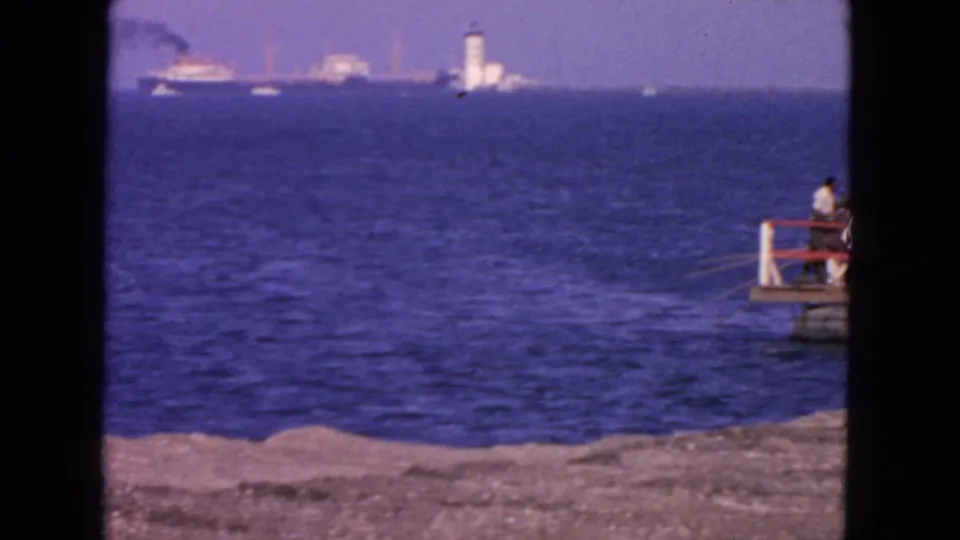 1963 San Francisco coastal view with blue water, distant ships, a white tower-like lighthouse, shoreline, and a person seated on a small pier.