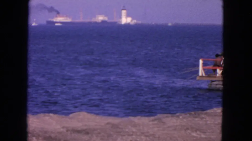 1963 8mm coastal scene in San Francisco with waves in the foreground, distant harbor structures, ships, smoke, and a pier observer.