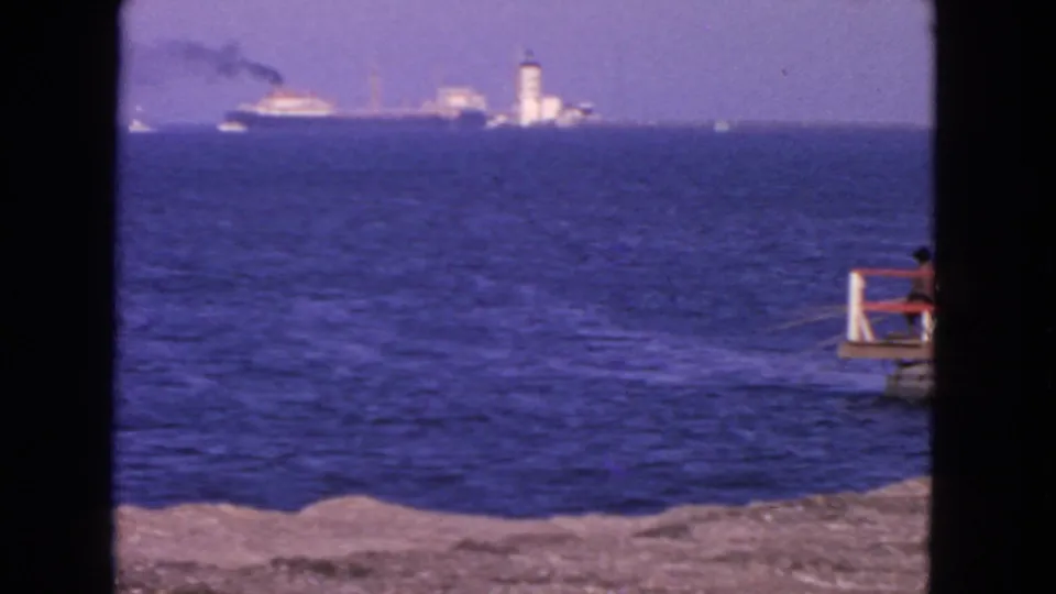 San Francisco 1963 home-movie frame showing blue water, distant ships, a white tower-like lighthouse, smoke, shoreline, and a pier at right.
