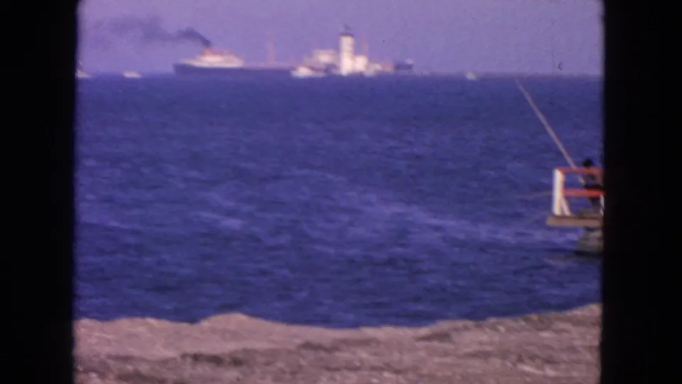 San Francisco coastal home-movie frame with deep blue water, a distant ship and tower, and the edge of a small pier with an angler at right.