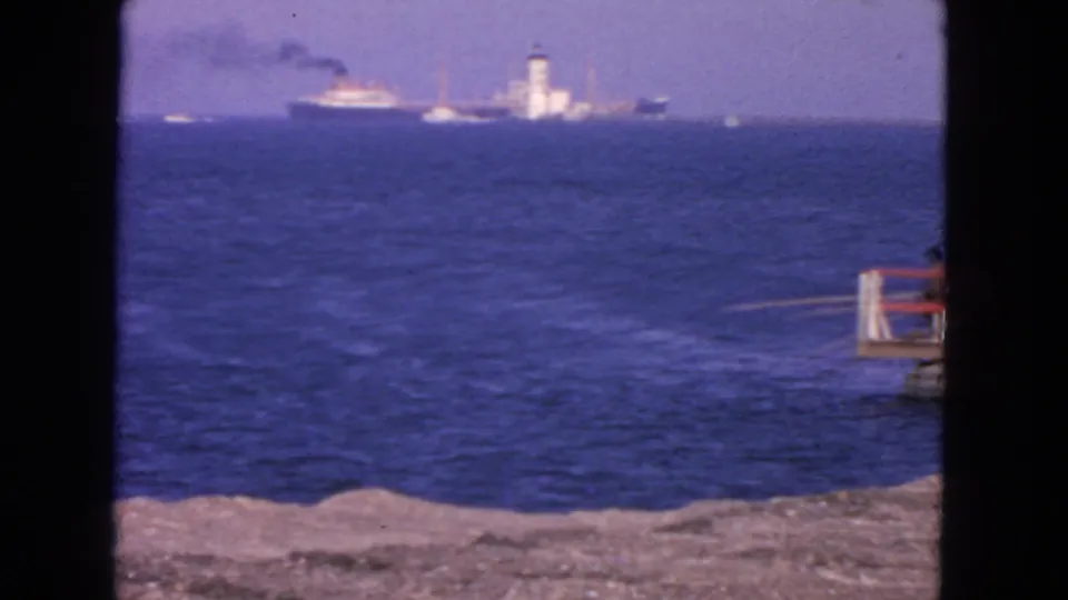 1963 San Francisco shoreline view with rocky foreground, blue ocean, a distant smoking ship, and a lighthouse-like tower on the horizon.