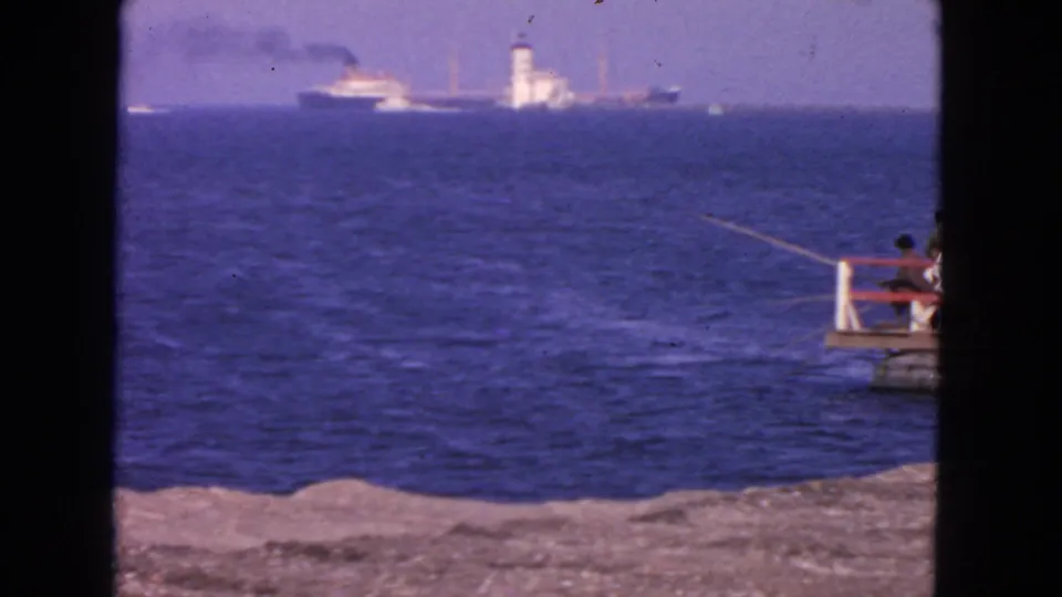 San Francisco coastal frame from 1963 showing people fishing from a small pier at right, with a distant ship and tower across the water.