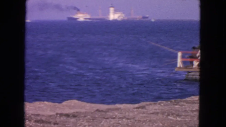 1963 San Francisco home-movie frame showing blue ocean, rocky shoreline, a distant ship and tower, with a pier edge at right.