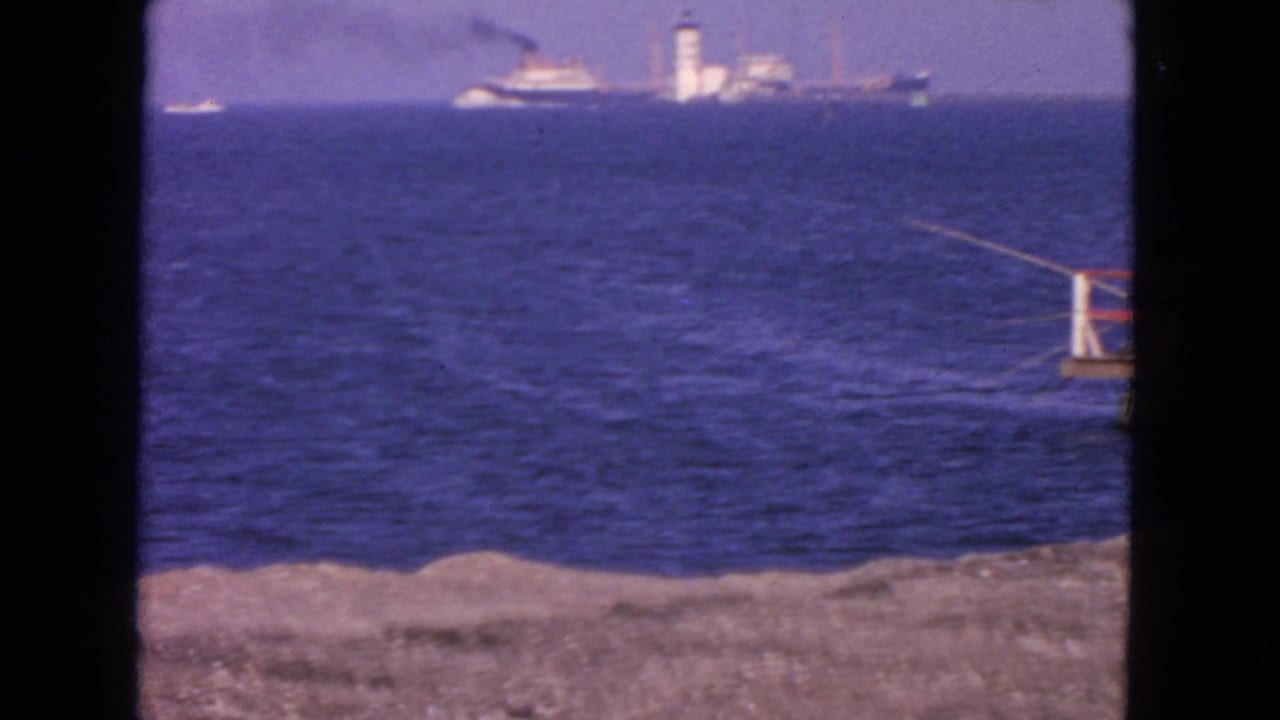 1963 San Francisco waterfront frame with a passenger ship and tower on the horizon, blue water, and a cropped pier railing.