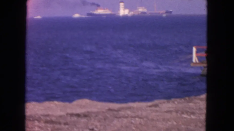 Soft 8mm frame of the San Francisco shoreline with blue ocean, rocky foreground, distant ship, and tower on the horizon.