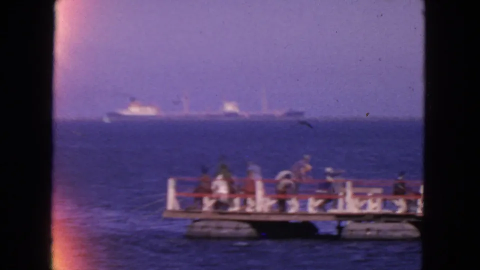 1963 San Francisco home-movie frame showing people on a small waterfront pier, blue ocean, and distant ships on the horizon.