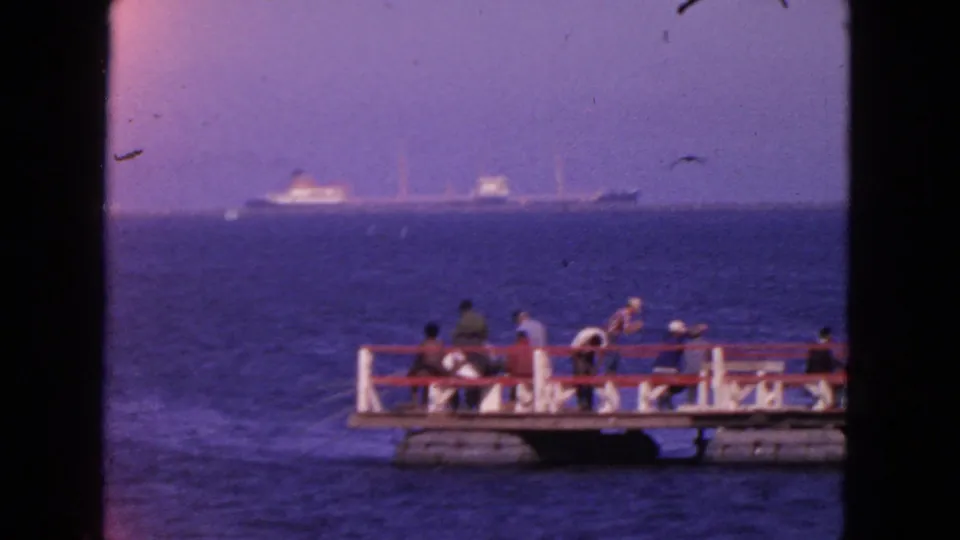 1963 San Francisco home-movie frame showing visitors on a small waterfront dock with distant cargo ships across the bay.