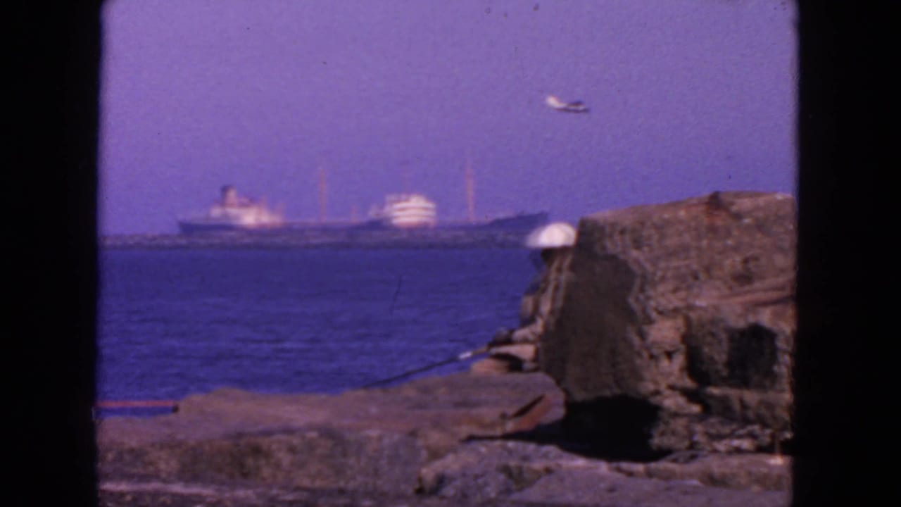 Seated person in a white hat fishing near rocks with ships across the water.