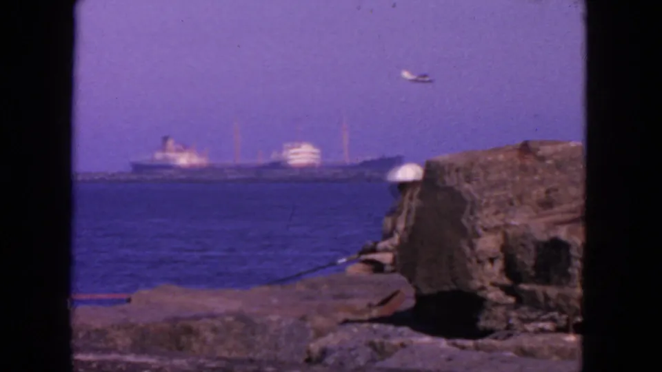 1963 San Francisco waterfront frame with a seated fisherman near a rocky pier edge and large ships in the distance.