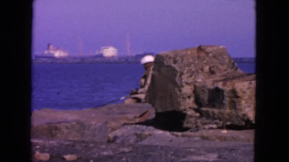 Home-movie view of a rocky San Francisco waterfront area, a seated fisherman, and ships blurred on the far shore.