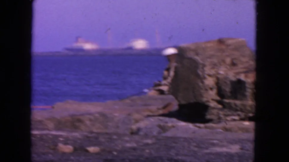 1963 waterfront frame showing a fisherman in a white hat seated beside rocks, with deep blue bay water and distant shipping.