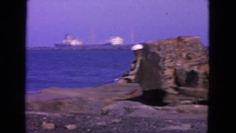 1963 San Francisco home-movie frame showing a person in a white hat seated beside a stone ledge with ships far across the bay.