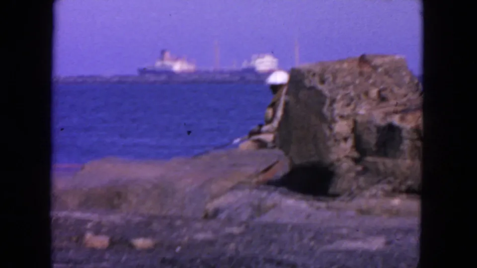 1963 San Francisco coastal frame with a white-hatted figure by rough stonework and ships crossing the blue bay in the background.