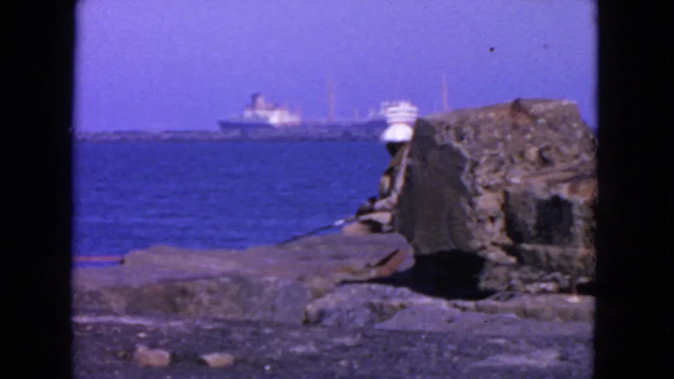 San Francisco 1963 8mm frame showing bay traffic beyond a rocky ledge, with a seated person in a white hat near the shoreline.
