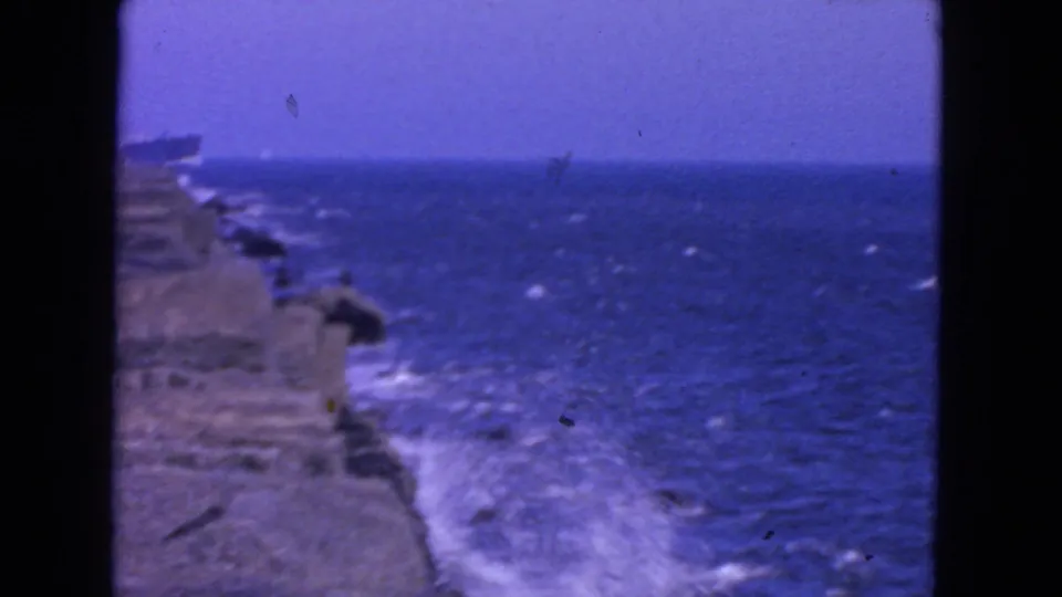 1963 San Francisco 8mm frame showing Pacific waves breaking against a rocky shoreline with a distant horizon and soft aged-film color.