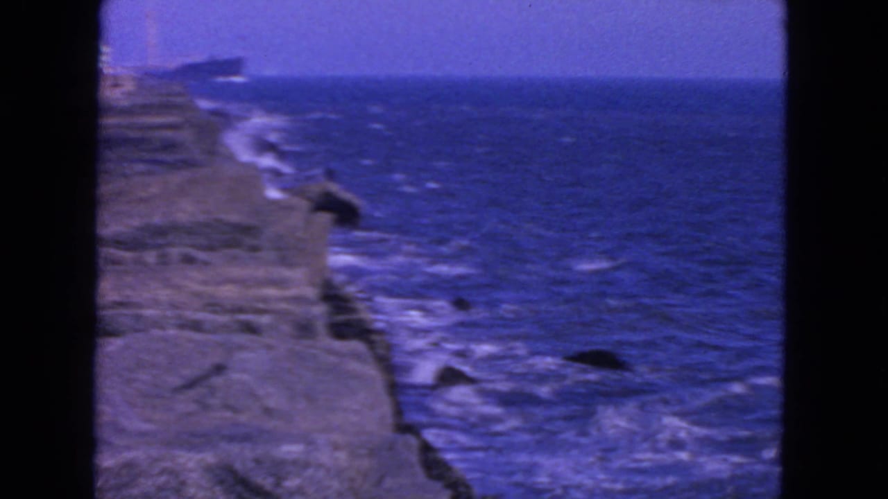 Rocky coast and blue ocean with white surf and a distant ship near the horizon.