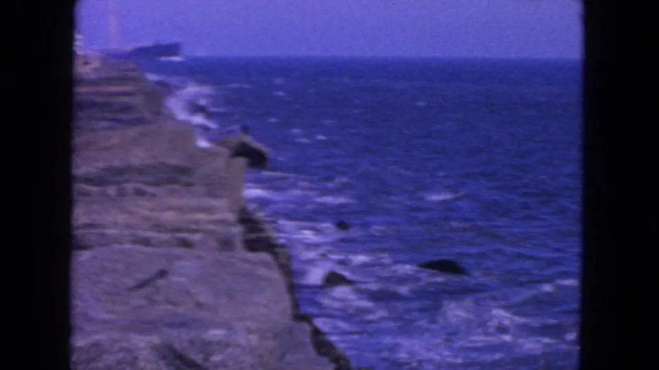 1963 San Francisco coastal view with waves striking rocks and a large ship faintly visible offshore under a blue horizon.