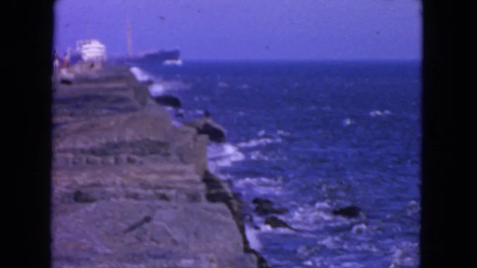 1963 San Francisco 8mm view of waves below a rocky coastal ledge, with small visitors faintly visible near the top of the frame.