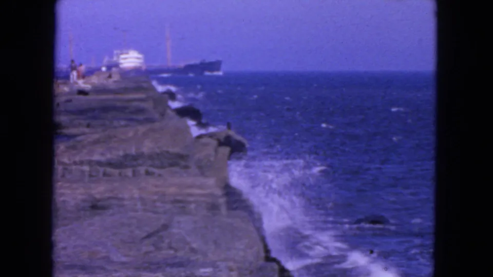 1963 San Francisco home-movie frame showing visitors on a rocky shoreline, surf below, and a large ship in the distance.
