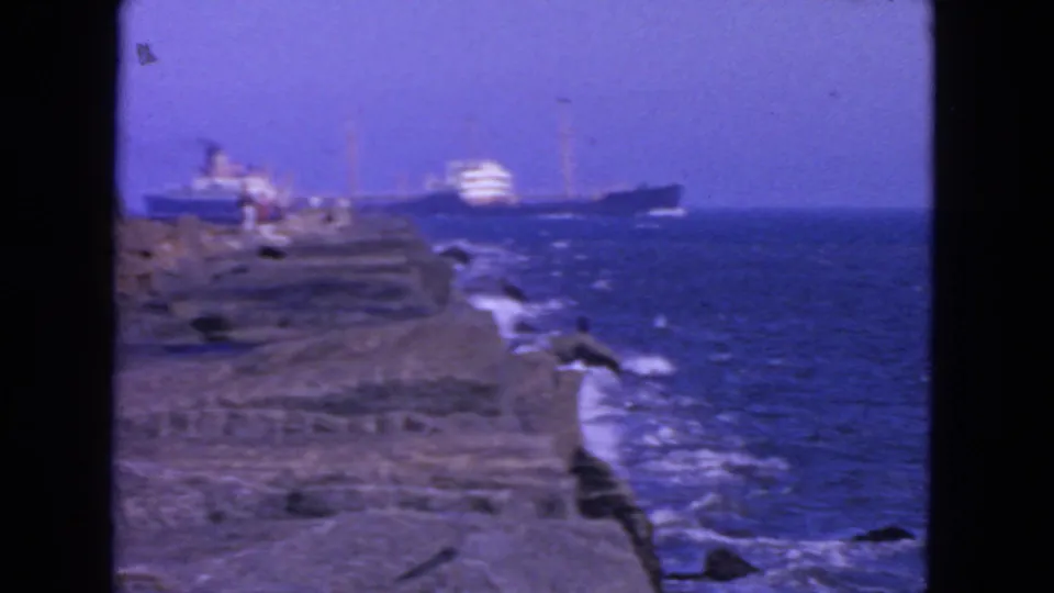 1963 San Francisco home-movie frame with surf, layered rocks, tiny people, and ships offshore.