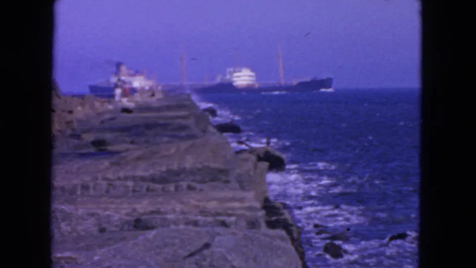 1963 San Francisco seaside frame with waves along rocks, tiny jetty figures, and offshore vessels.