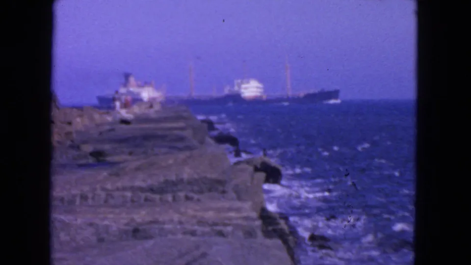 1963 San Francisco home-movie frame showing a large ship at sea beyond a rocky coastal edge with small distant people nearby.