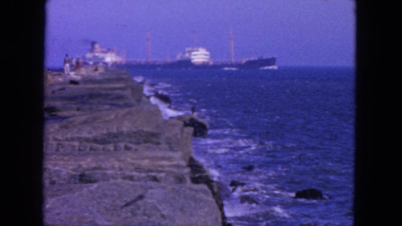1963 San Francisco coastal frame with a large ship offshore, waves against dark rocks, and small distant figures near the ledge.