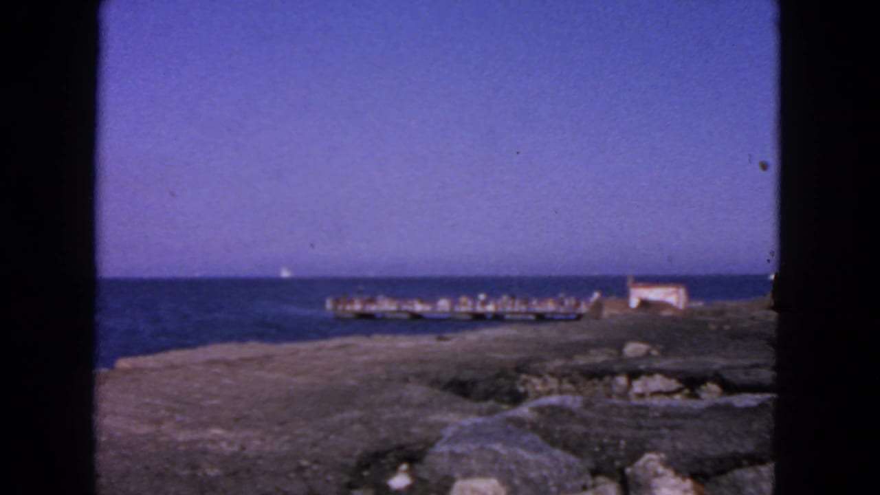 Pier and small waterside structure seen across blue water with rocky ground in front.