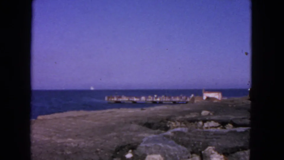 1963 San Francisco waterfront view with a pier, small white building, rocky foreground, and calm horizon.