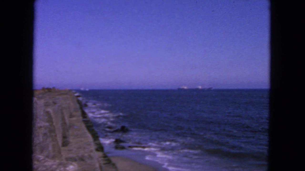 Concrete breakwater beside blue ocean with white surf and a distant ship.