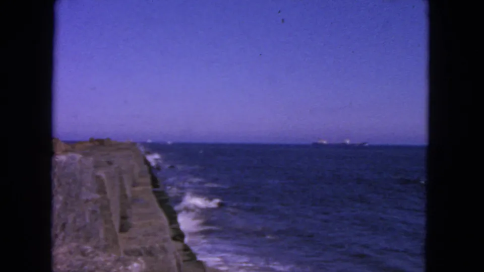 Home-movie frame from 1963 San Francisco showing ocean waves breaking beside a concrete breakwater under a clear sky.