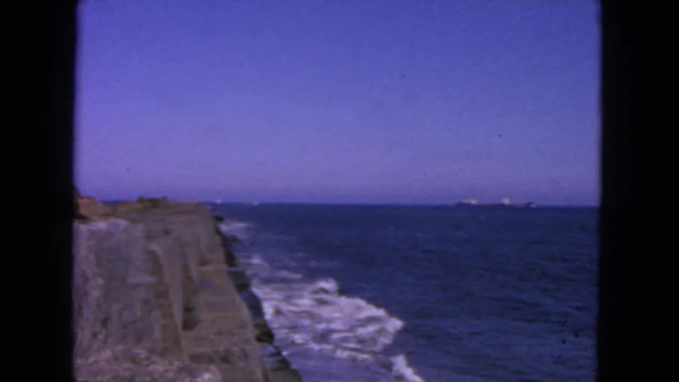 1963 San Francisco coastal frame with white surf, blue ocean, concrete breakwater, and a faint ship near the horizon.