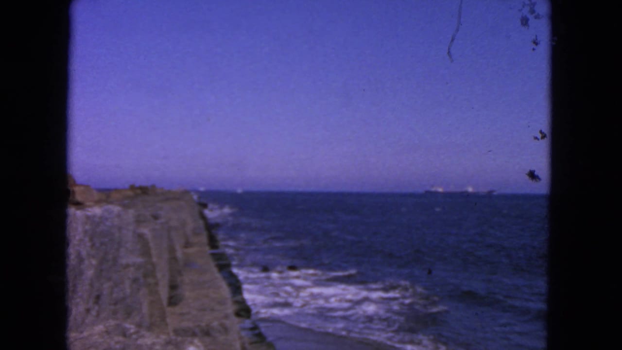 Stone pier beside waves with open ocean and a distant ship on the horizon.