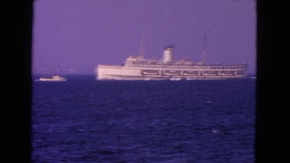Closer 1963 San Francisco view of a large white passenger ship on blue water, with a small boat alongside and hazy sky behind it.
