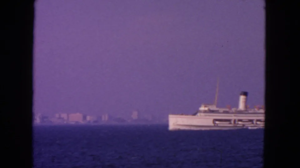 1963 San Francisco-area 8mm frame of a white passenger ship near the right edge, with distant shore structures softened by haze.