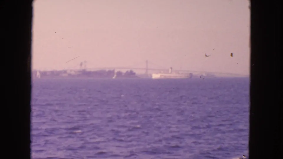 1963 San Francisco Bay view with a distant ship near a suspension bridge, seen across textured water in faded 8mm color.