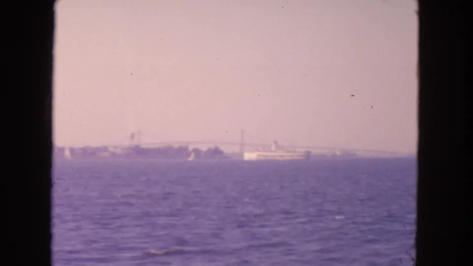1963 San Francisco Bay frame showing a ship, bridge span, low shoreline, and hazy water through a home-movie film border.
