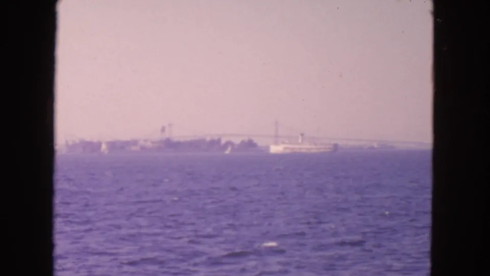 1963 San Francisco Bay home-movie frame with a distant ship, suspension bridge, and rippled water under a pale hazy sky.