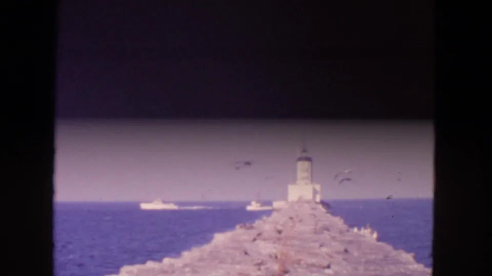 1963 San Francisco Bay view of a stone breakwater leading to a small lighthouse, with boats and seagulls offshore.