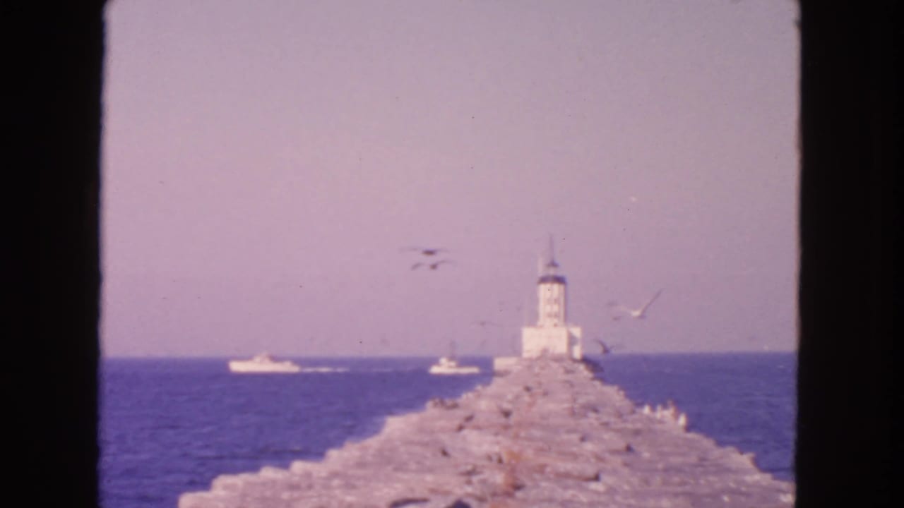 Stone breakwater leading to a white lighthouse with gulls and boats over blue water.