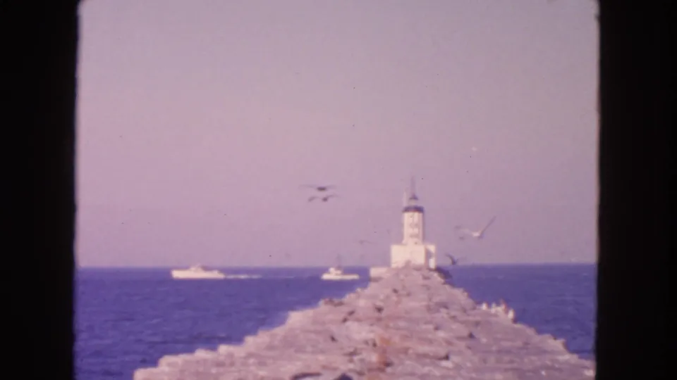 1963 San Francisco coastal 8mm frame with a stone breakwater, white lighthouse, gulls in flight, and small boats offshore.