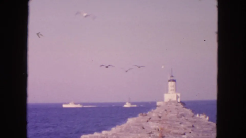Faded 8mm San Francisco coastal frame showing a lighthouse, breakwater, gulls, and small boats against the bay horizon.