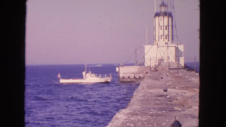 1963 San Francisco home-movie frame of a boat near a white coastal lighthouse, with the rocky pier filling the right side.