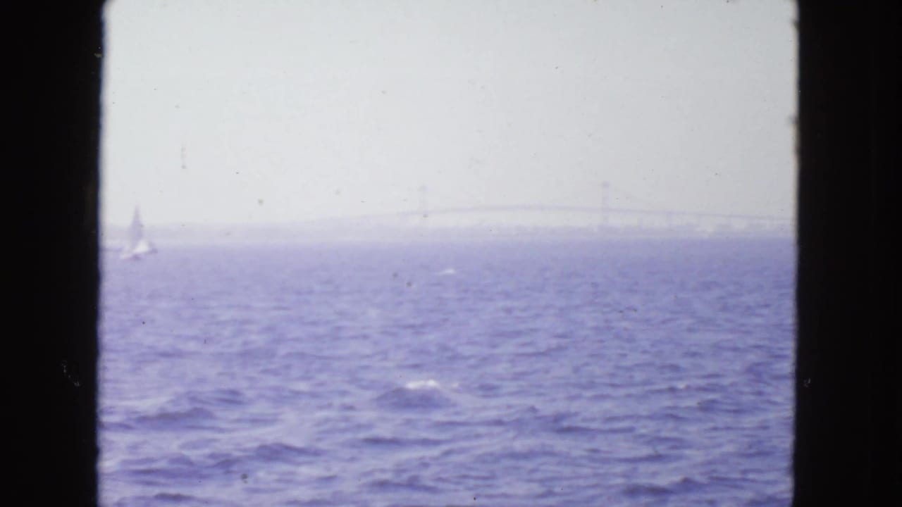 Choppy bay water with a faint bridge on the horizon and a small sailboat at left.