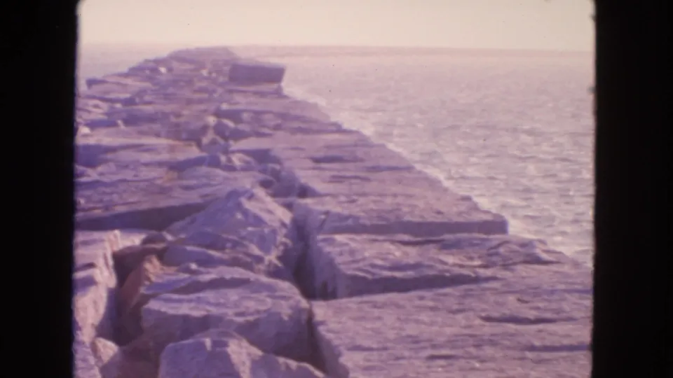 San Francisco-area coastal jetty in 1963 home-movie footage, with large rocks, surrounding bay water, and haze in the distance.