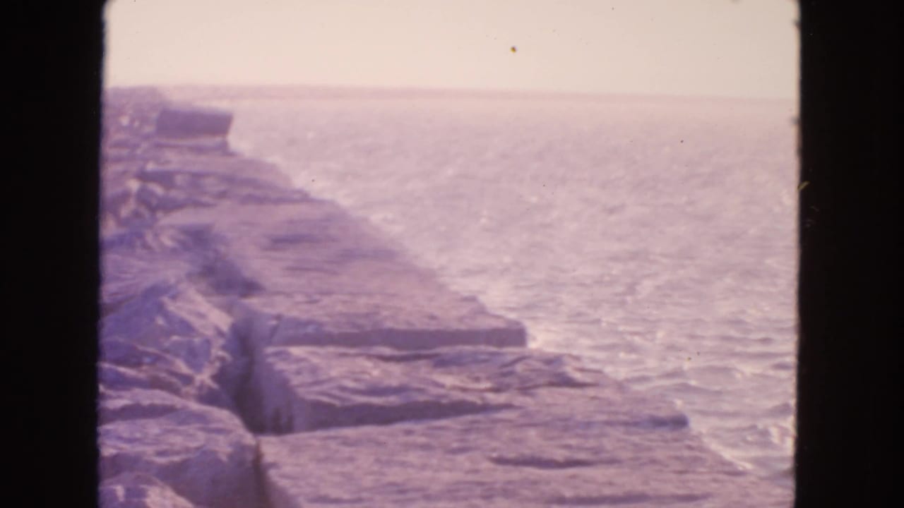 1963 8mm San Francisco coastal view with layered stone breakwater, calm ocean, haze, and dark frame edges.