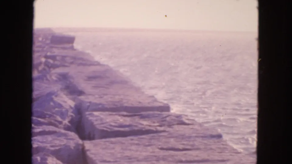 1963 8mm San Francisco coastal view with layered stone breakwater, calm ocean, haze, and dark frame edges.