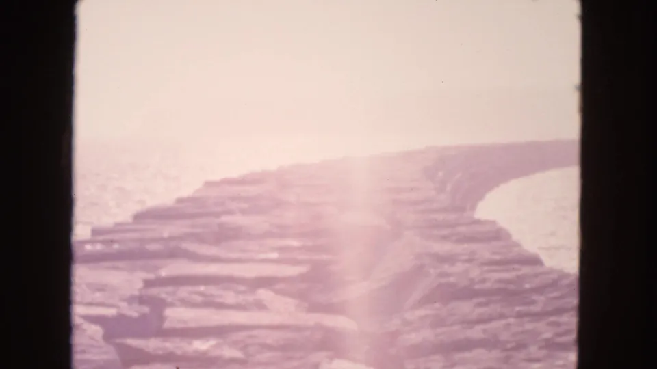 1963 8mm coastal frame showing layered stone slabs, ocean glare, haze, and a pale vertical exposure flare.