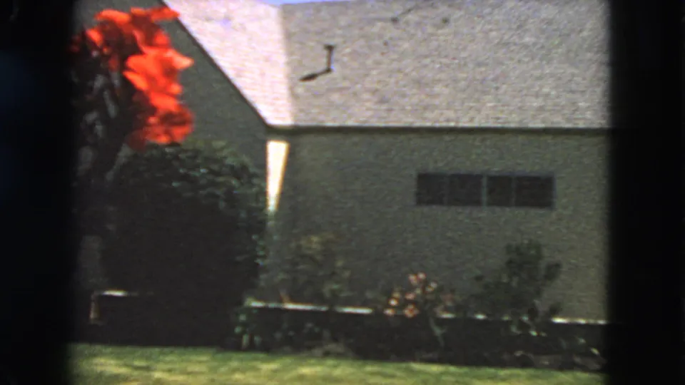 A static 1963 San Francisco home-movie view shows red flowers, lawn, shrubs, and a textured house wall with small windows.